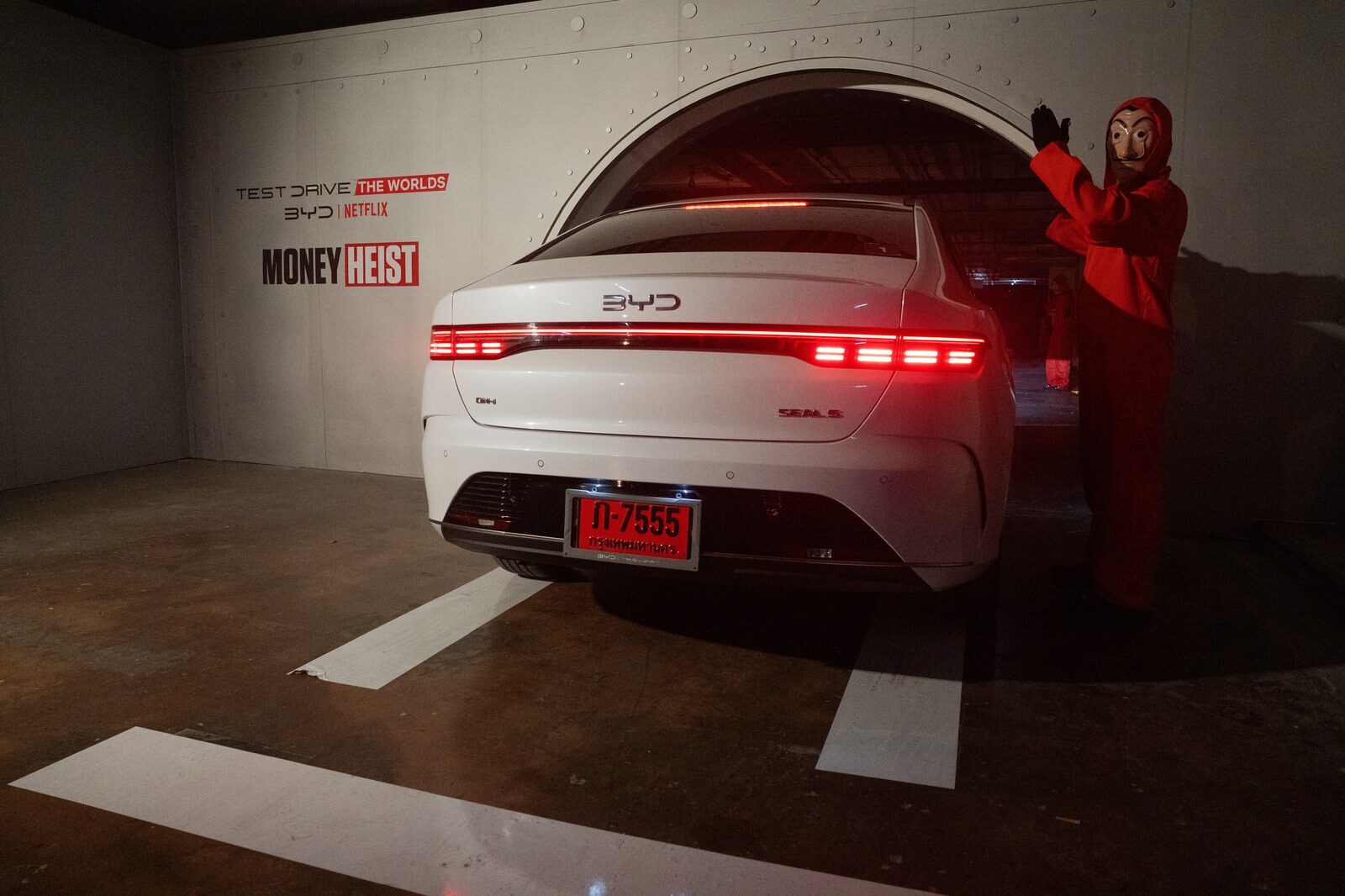 BYD สีขาวมองจากด้านหลังในโซน Money Heist มีป้าย BYD x Netflix และผู้สวมชุดแดงหน้ากากยืนข้างรถ