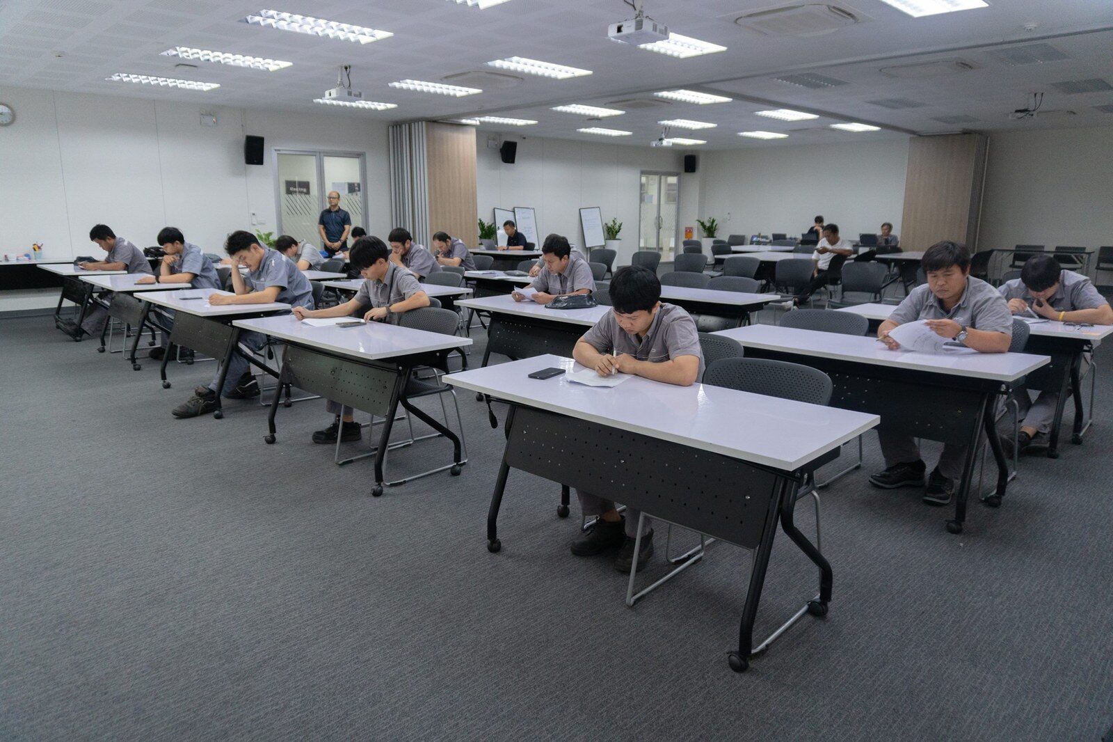ผู้เข้าแข่งขันในชุดช่างนั่งทำข้อสอบข้อเขียนในห้องสอบขนาดใหญ่