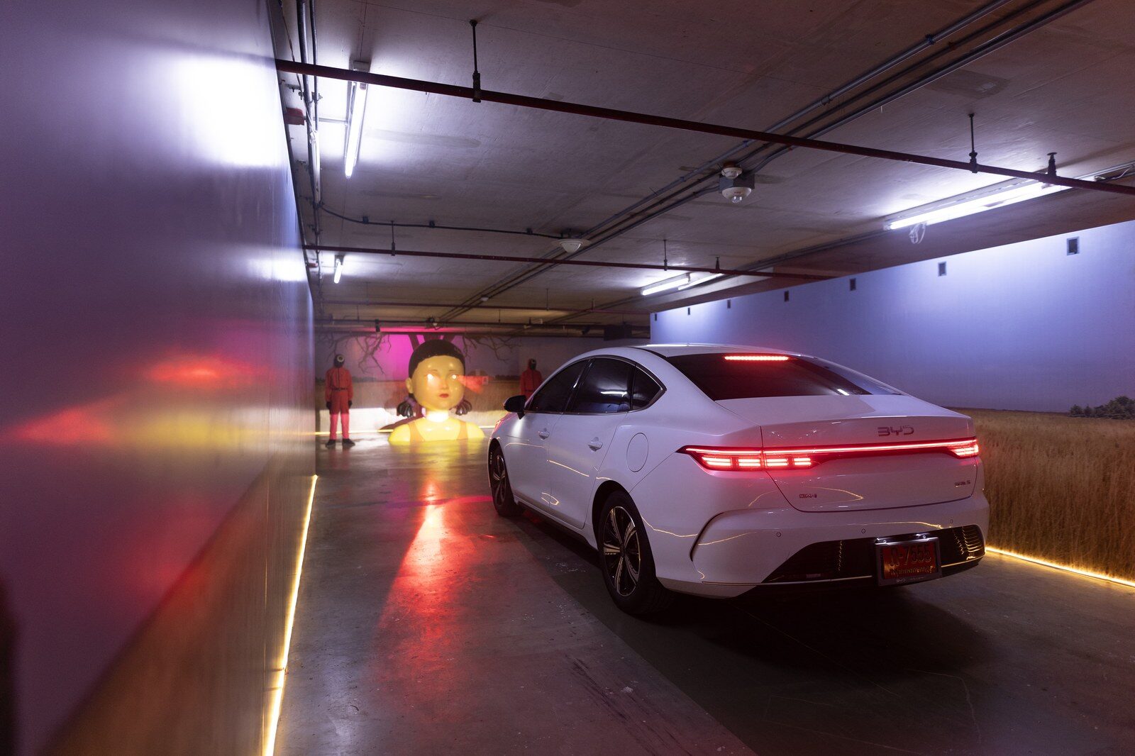 BYD ซีดานสีขาวมองจากด้านหลัง จอดในทางเดินที่มีหุ่นตุ๊กตาขนาดใหญ่และไฟแถบขอบพื้น