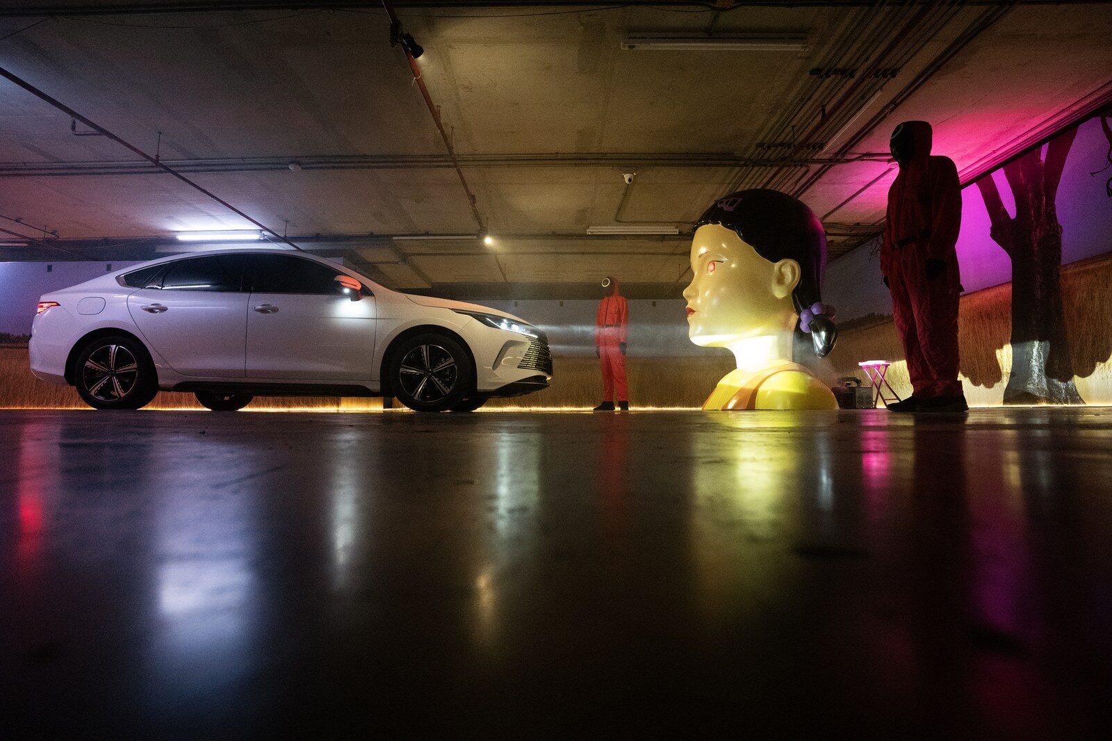 BYD ซีดานสีขาวด้านข้างจอดอยู่ใกล้หุ่นตุ๊กตาขนาดใหญ่และผู้สวมชุดแดงในลานจอดรถ