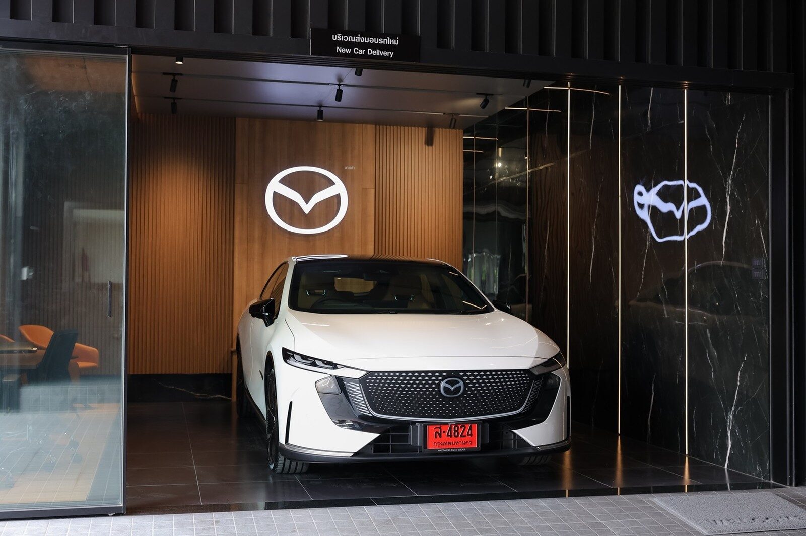รถมาสด้าสีขาวจอดในโซนส่งมอบรถใหม่ (New Car Delivery) ที่มีโลโก้มาสด้าบนผนังไม้