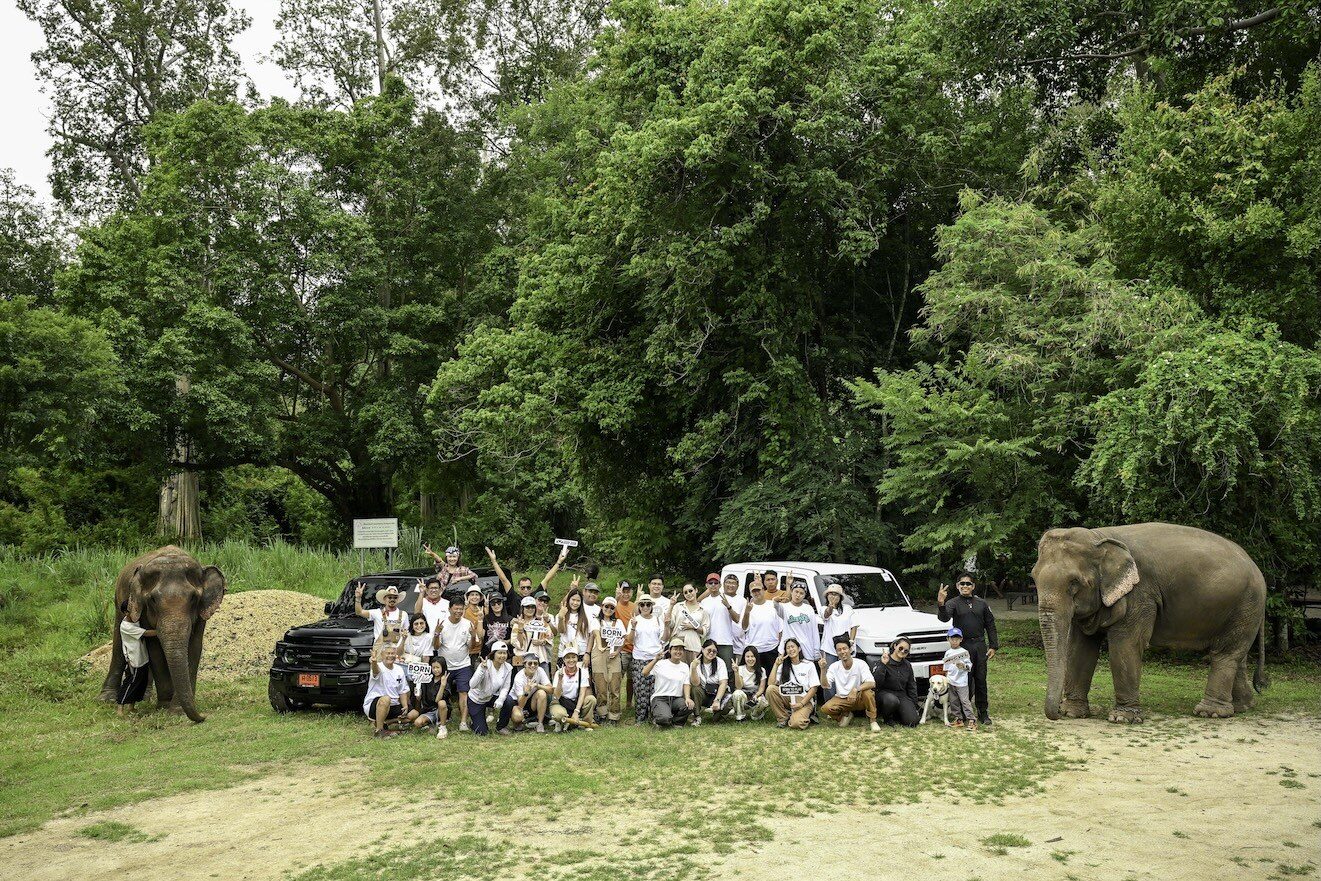 ผู้ร่วมกิจกรรมถ่ายภาพหมู่กับช้างสองเชือกและรถ SUV สองคันกลางป่า