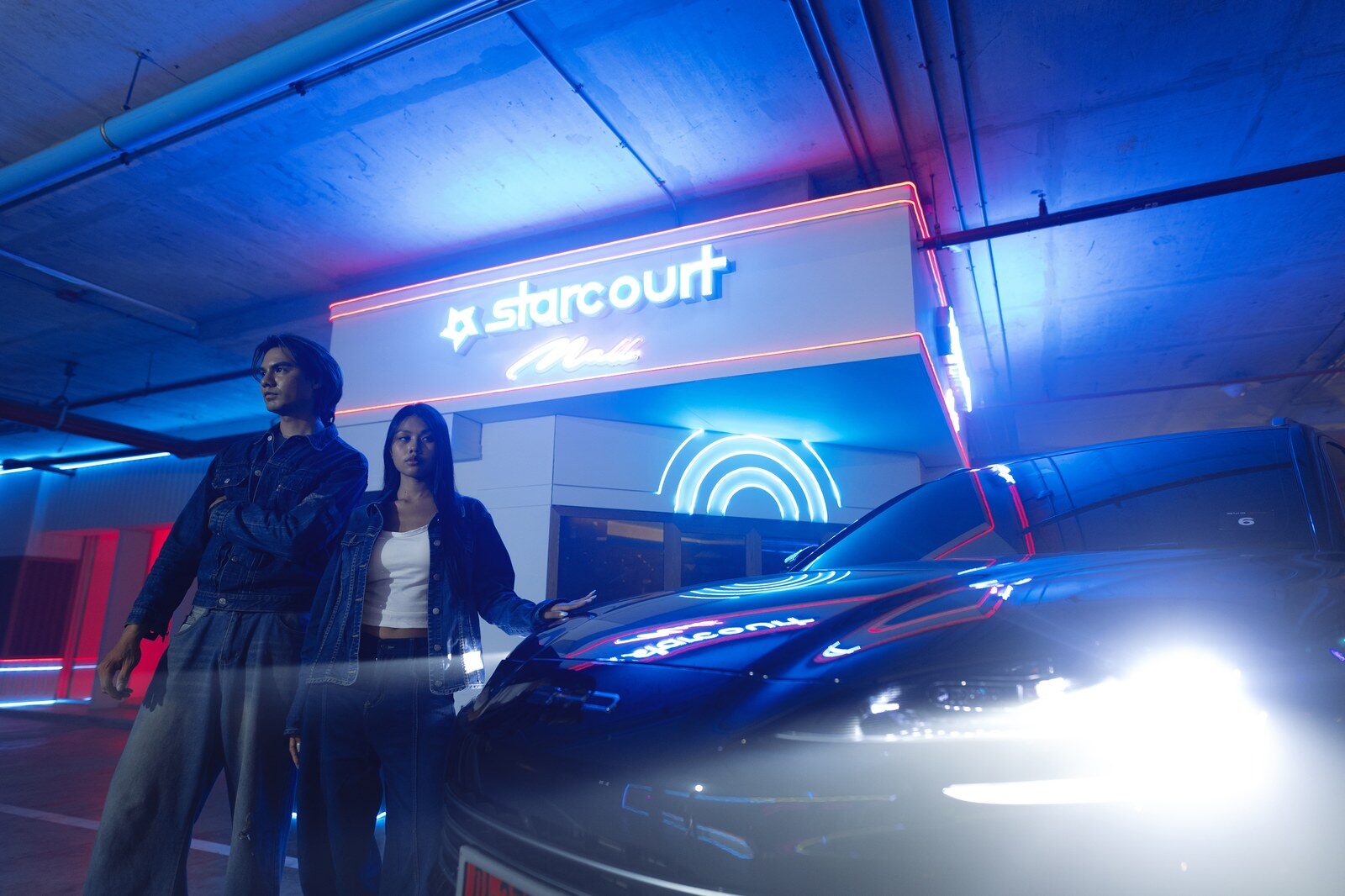 คู่ผู้ร่วมงานในแจ็คเก็ตยีนส์ยืนข้างรถ BYD หน้าฉากป้ายนีออน starcourt