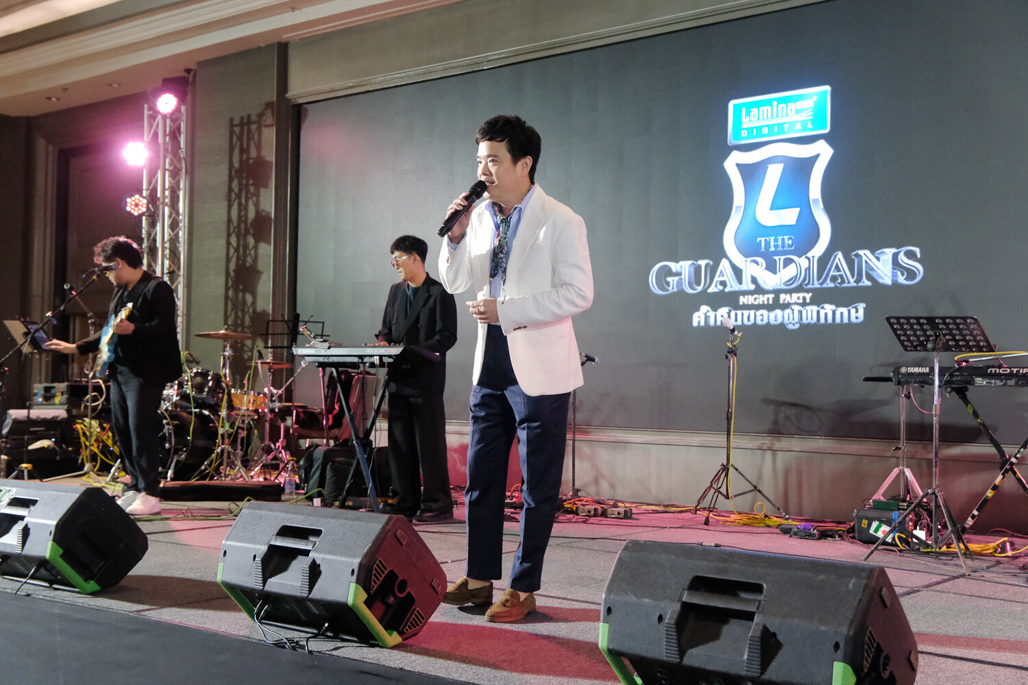 ศิลปินในสูทขาวถือไมค์ร้องเพลง มีวงดนตรีอยู่ด้านหลัง ฉากหลัง The Guardians Night Party